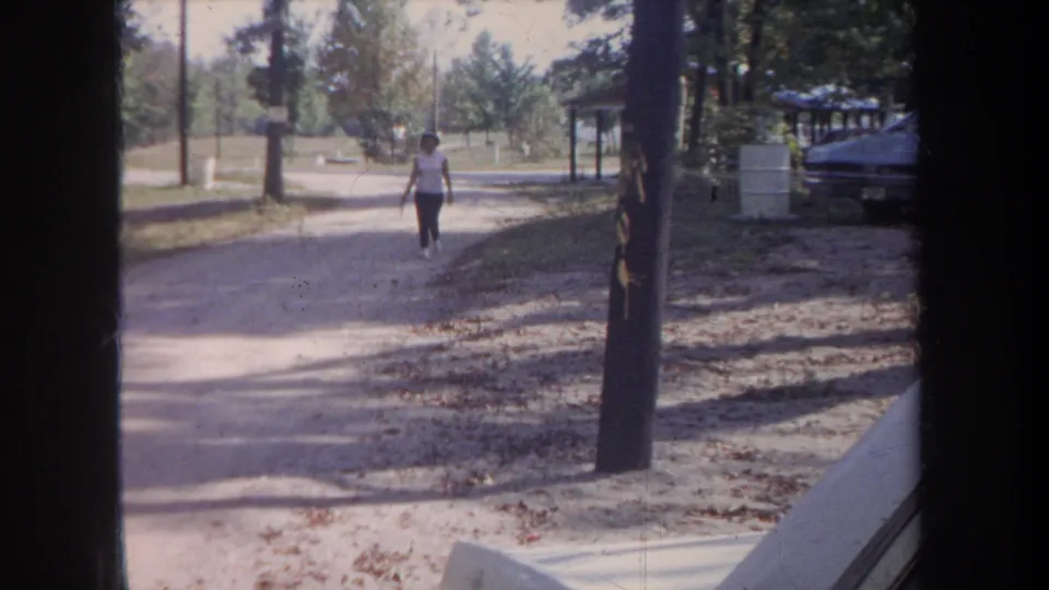 Near the end of the clip, the woman continues toward the camera on the gravel path, with trees, shadow, and a parked car at the edge of frame.