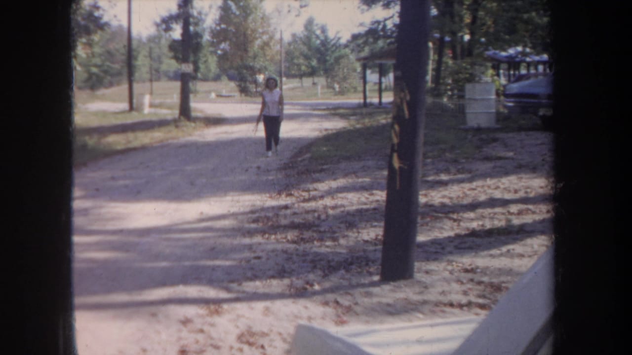 Woman walking on an unpaved road lined with trees, seen through a dark window frame.