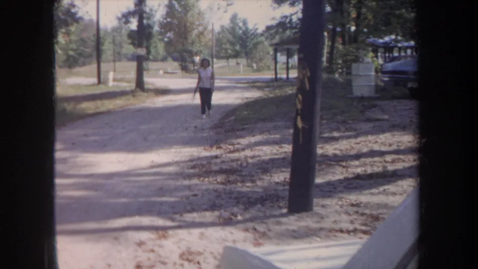 1968 Michigan home-movie frame showing a woman in a white top walking toward the camera on a wooded unpaved road.