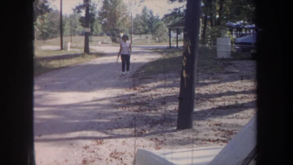 1968 Michigan 8mm frame of a woman walking alone on a sunlit rural road, with trees and roadside structures behind her.