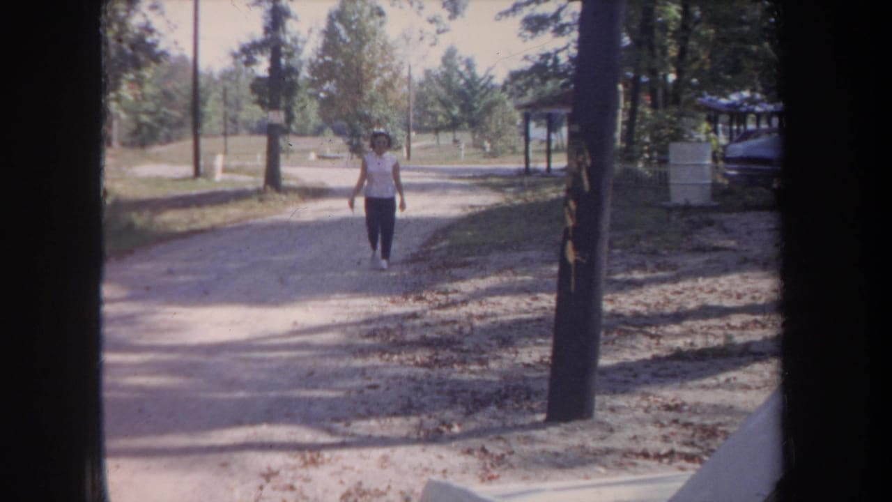 1968 Michigan home-movie still with a woman walking closer on an unpaved road through a wooded outdoor area.