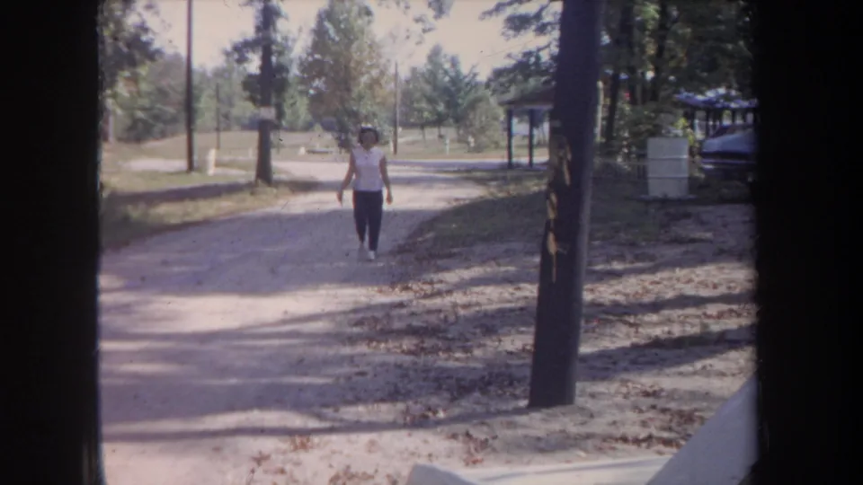 1968 Michigan home-movie still with a woman walking closer on an unpaved road through a wooded outdoor area.