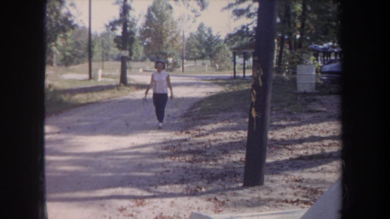 1968 Michigan frame showing the woman near the foreground on a rural unpaved road, filmed from a stationary position.