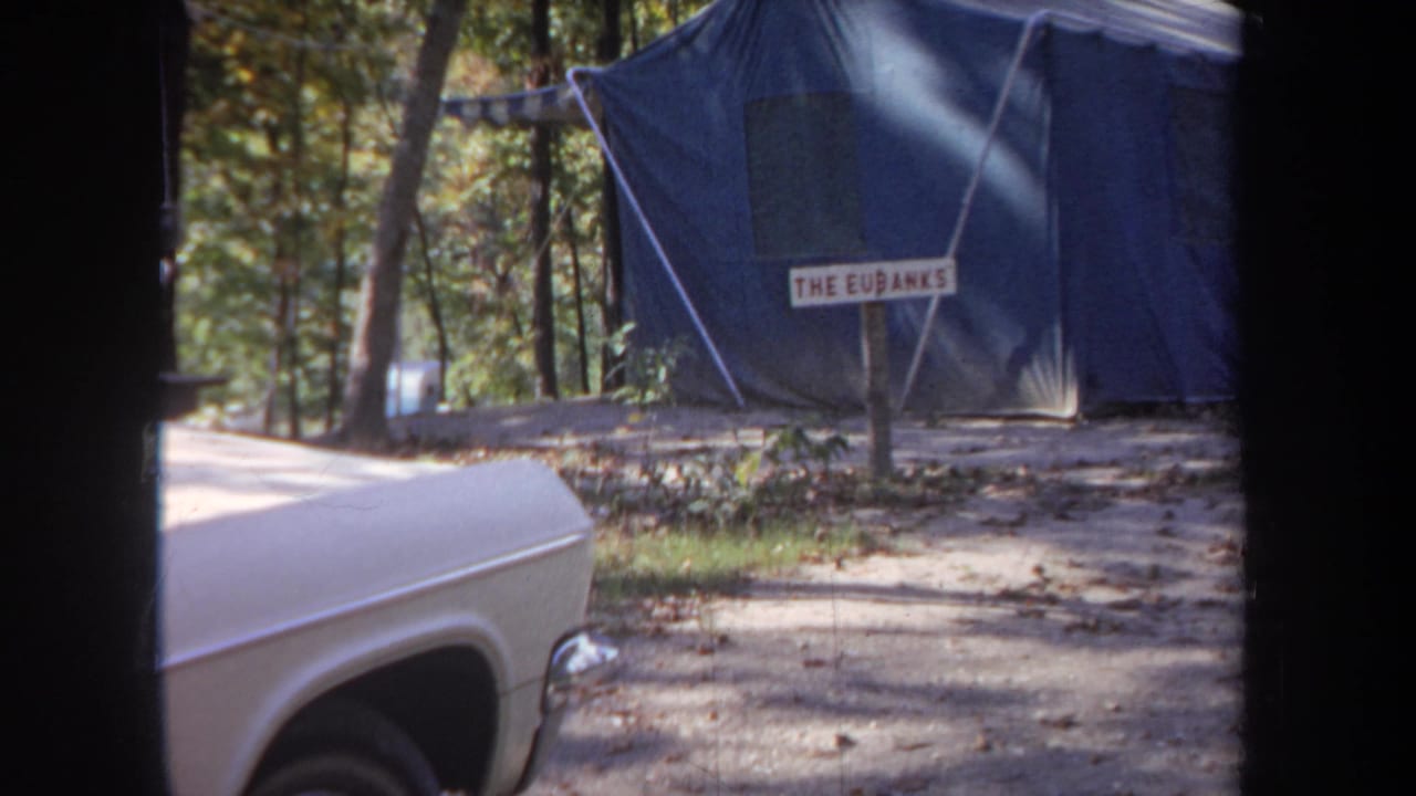 1968 Michigan home-movie frame showing a white car parked near a blue canvas tent marked with a family-name sign.