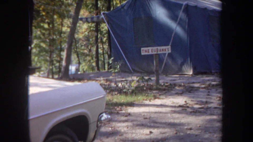 1968 Michigan home-movie frame showing a white car parked near a blue canvas tent marked with a family-name sign.