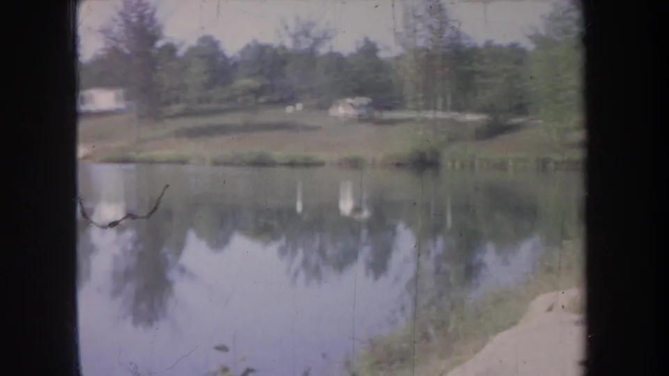 Michigan home-movie lake view with soft focus, tree reflections, grassy shoreline, and distant camping structures.