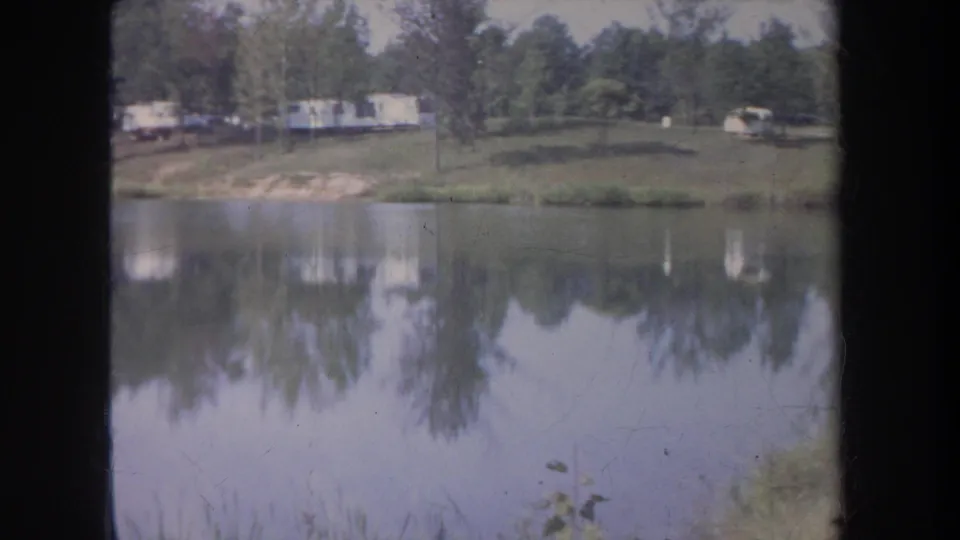 1968 Michigan lakeside frame showing calm water, reflected trees, and trailers or campers along the far bank.