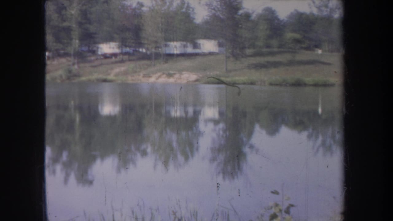 Calm lake with tree reflections and pale trailers on the far shore.