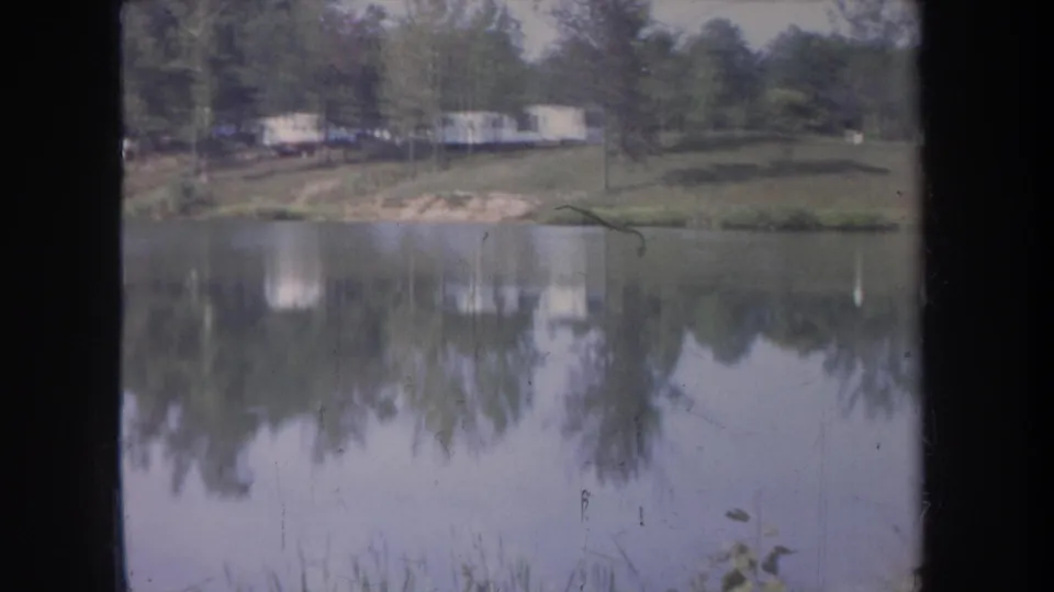 1968 Michigan 8mm frame showing a still lake, wooded shoreline, and distant pale trailers reflected in the water.