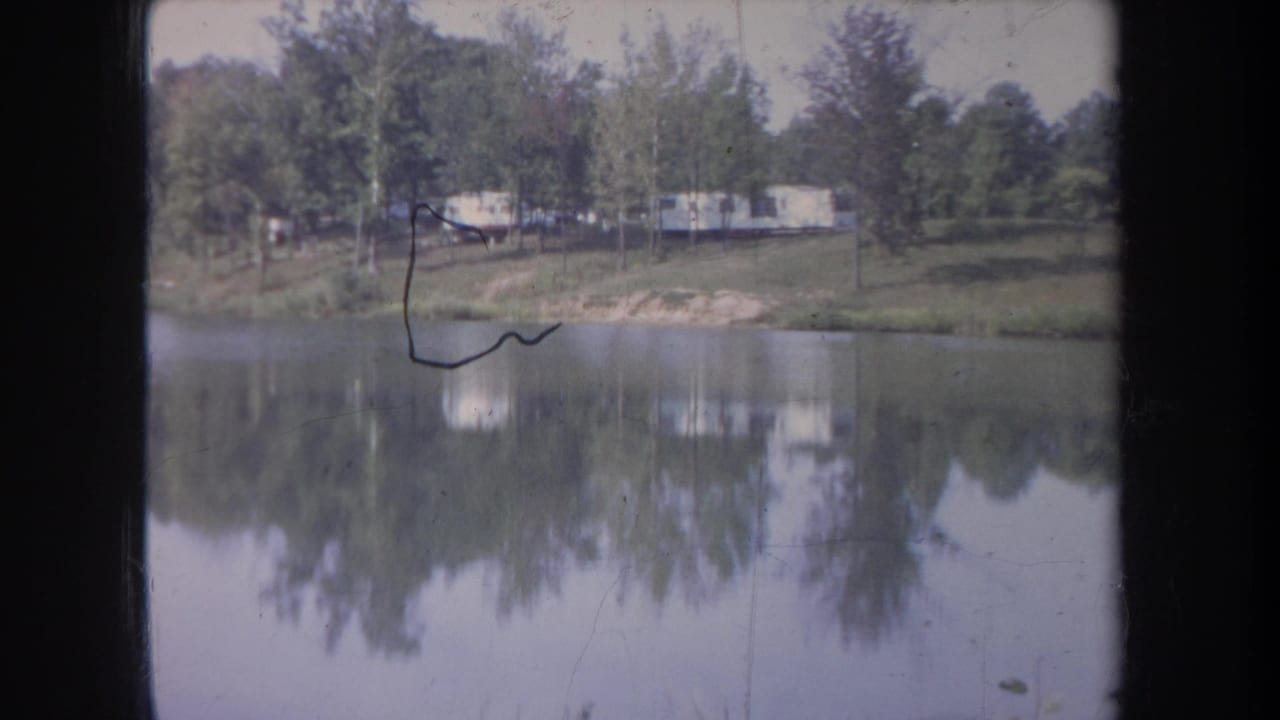 1968 Michigan 8mm frame with calm water, wooded shore, distant pale trailers, and visible film abrasion near the top.