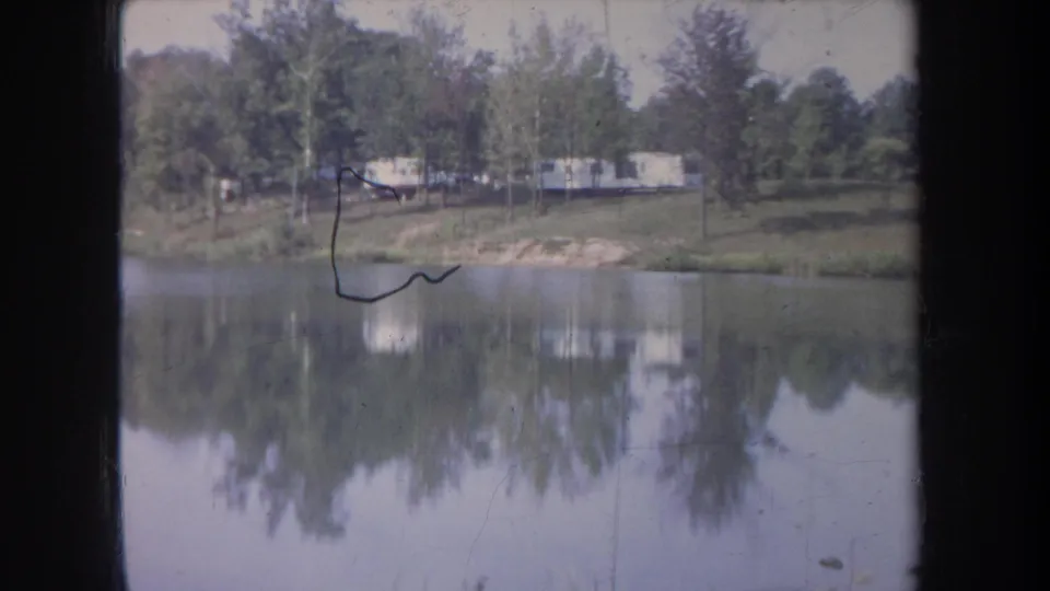 1968 Michigan 8mm frame with calm water, wooded shore, distant pale trailers, and visible film abrasion near the top.