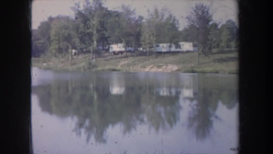 1968 Michigan 8mm frame showing a broad calm lake, tree-lined bank, pale trailers, and soft mirrored reflections.