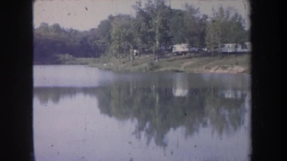 1968 Michigan 8mm frame showing a quiet lake bend, wooded shoreline, distant pale trailers, and muted reflections.