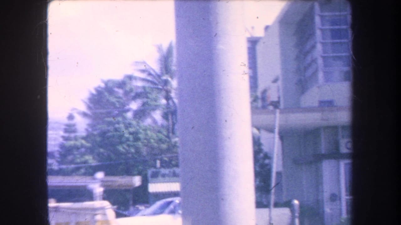A 1969 Hawaii street glimpse with palm trees, parked cars, a large column, and nearby commercial building edges.
