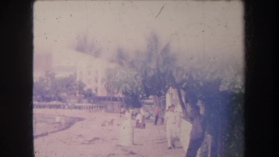 Faded 1969 Hawaii beachside view with pedestrians on a promenade, palm trees, sand, and a large shoreline building in the background.