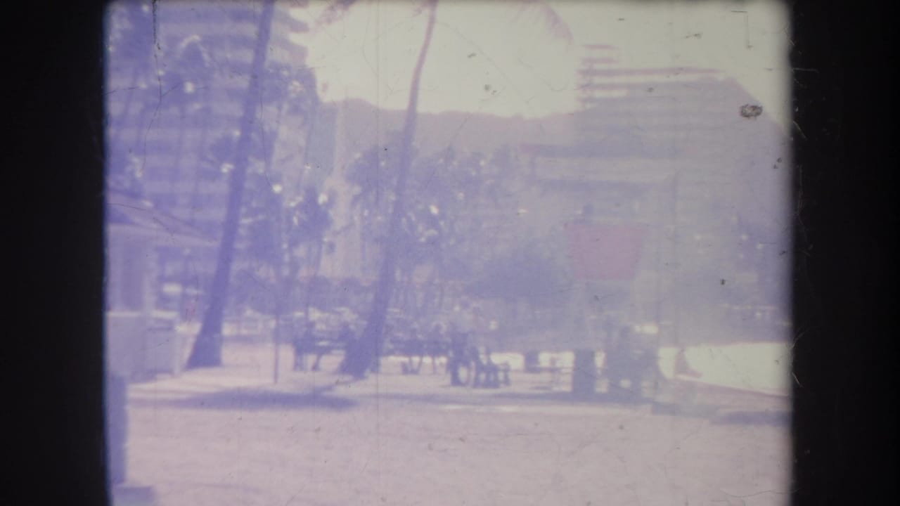 1969 Hawaii home-movie frame showing palms, a bright beach path, distant people, and high-rise buildings softened by purple film cast.