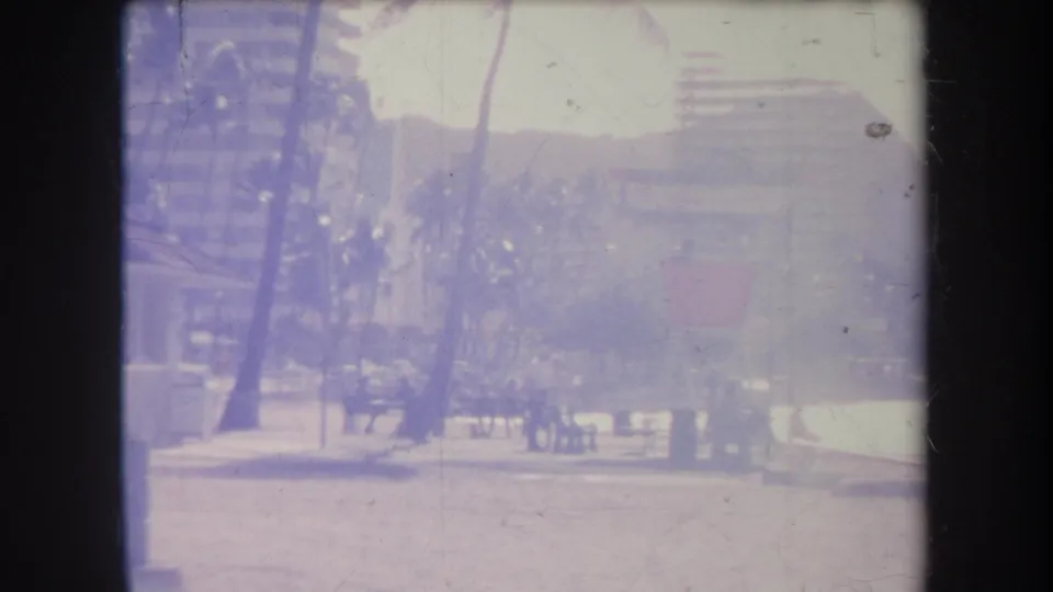 1969 Hawaii home-movie frame showing palms, a bright beach path, distant people, and high-rise buildings softened by purple film cast.