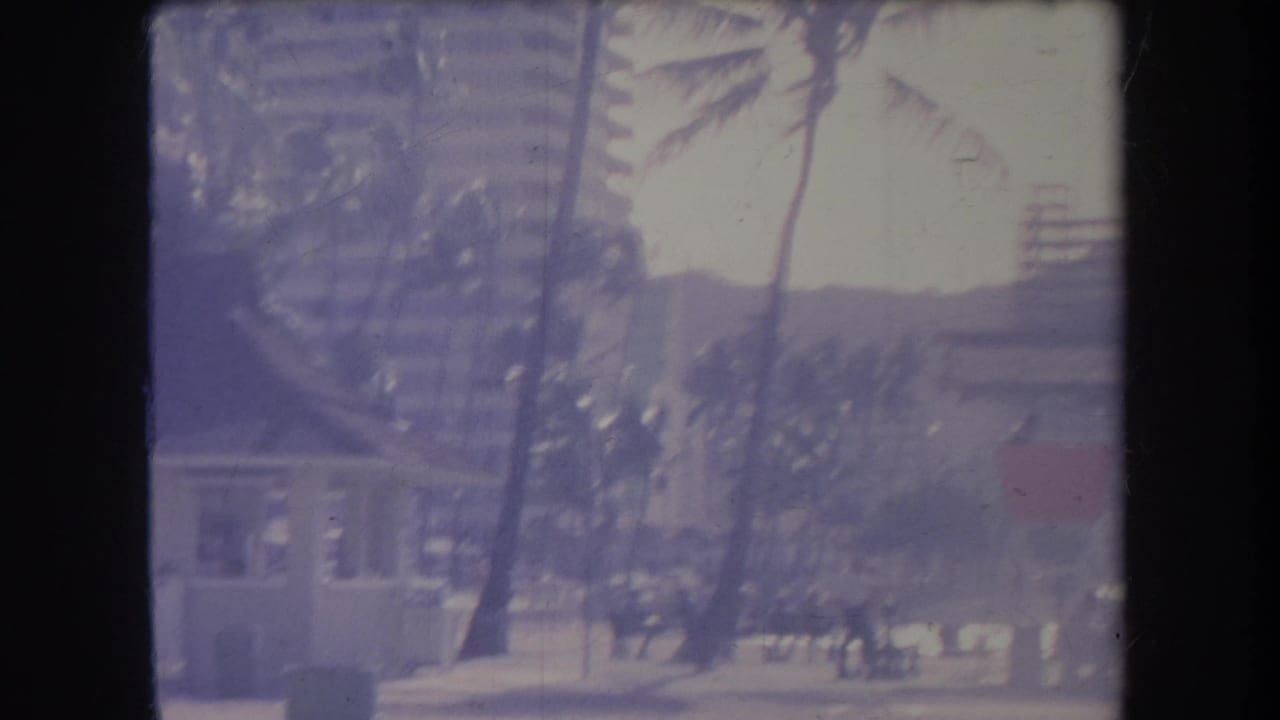 1969 Hawaii beach view with a lifeguard tower, palms, hotel-like buildings, and Diamond Head hillside beyond the shore.