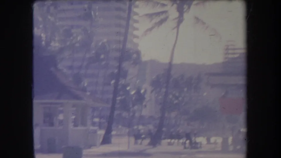 1969 Hawaii beach view with a lifeguard tower, palms, hotel-like buildings, and Diamond Head hillside beyond the shore.