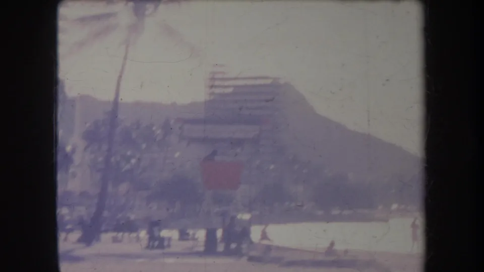 1969 Hawaii frame showing palms, a beach edge, distant people, a high-rise building, and Diamond Head softened by home-movie exposure.