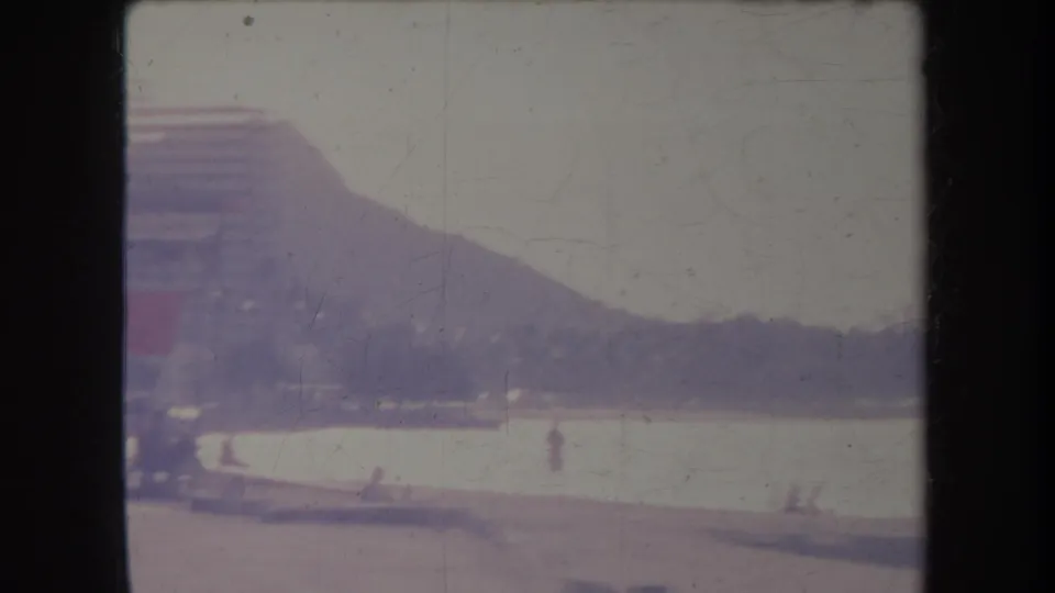 1969 Hawaii home-movie view across the beach toward Diamond Head, with small distant people near the water and a building at left.