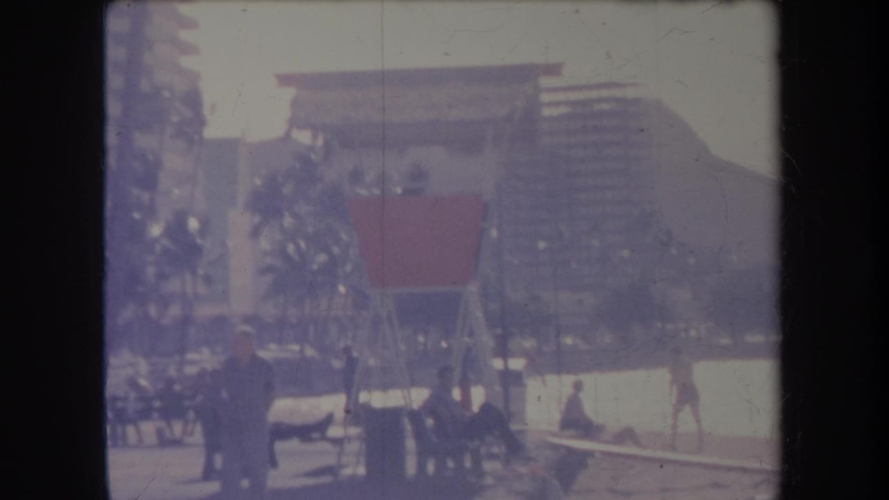 1969 Hawaii home-movie frame showing beachgoers near palms and a lifeguard-style structure, with tall buildings behind the shoreline.