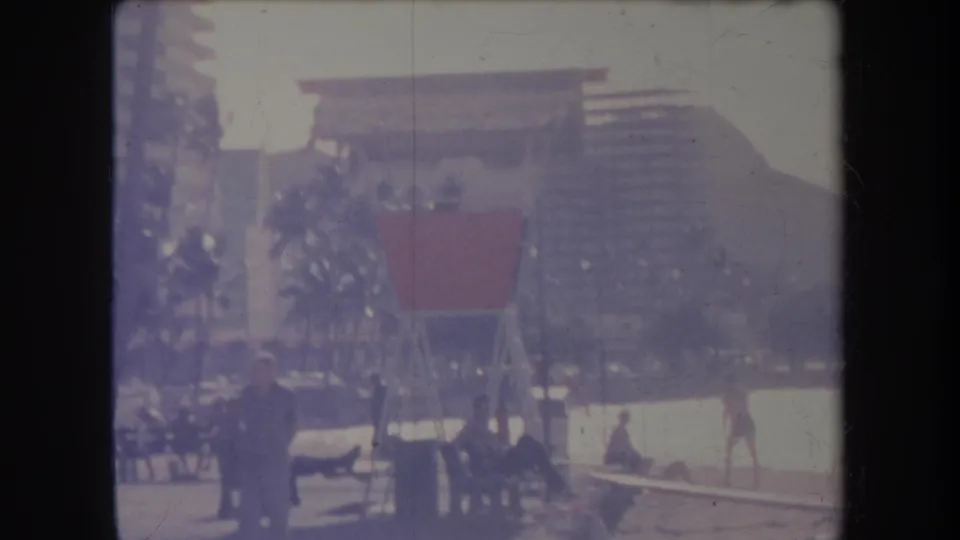 1969 Hawaii home-movie frame showing beachgoers near palms and a lifeguard-style structure, with tall buildings behind the shoreline.
