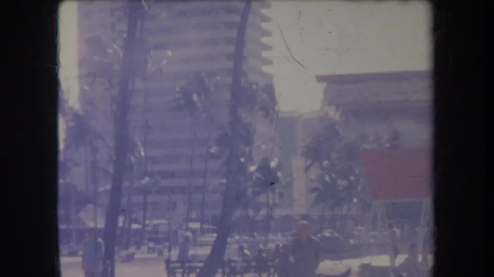 1969 Hawaii beach frame with people near palms, a tall modern building, and a red-roofed or colored beach structure in soft faded color.