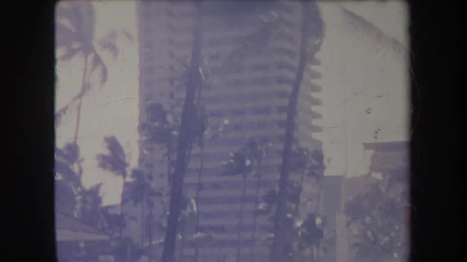 1969 Hawaii home-movie view of a tall waterfront building framed by palms, with beach activity partially visible below.