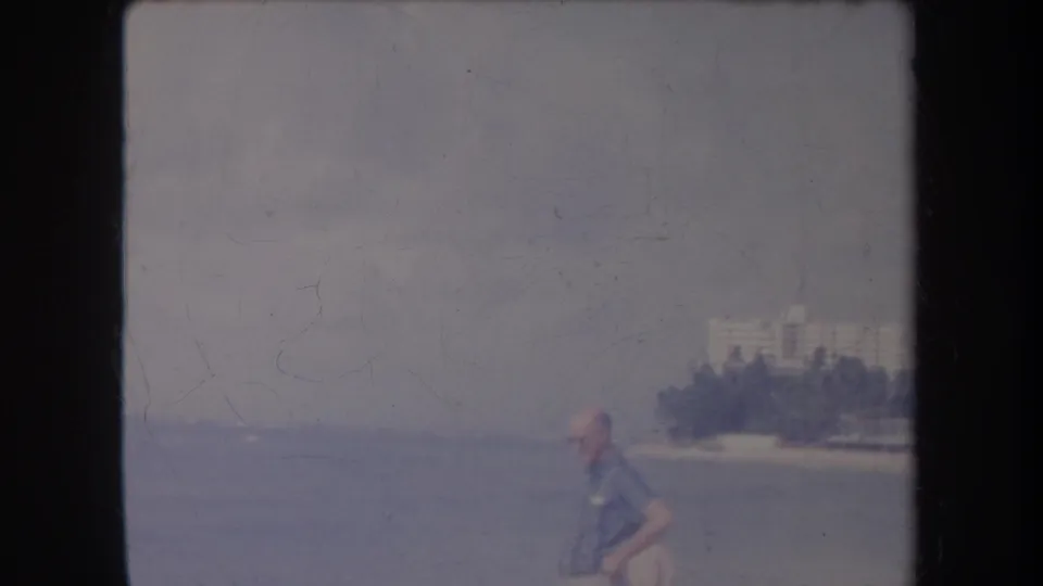 1969 Hawaii shoreline frame showing a man walking by the water, with distant palms and buildings along the coast.