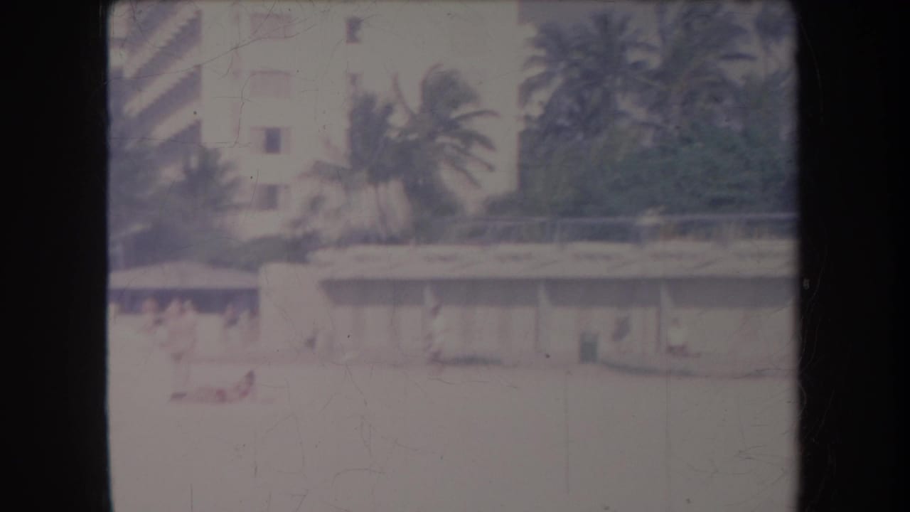 1969 Hawaii home-movie view of a bright beachfront area with palms, a low modern building, and small figures on the sand.