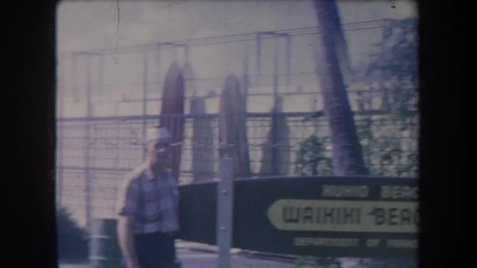 1969 Hawaii frame of a man in a hat near a Waikiki Beach Park sign, with surfboards behind fencing and the ocean in the background.