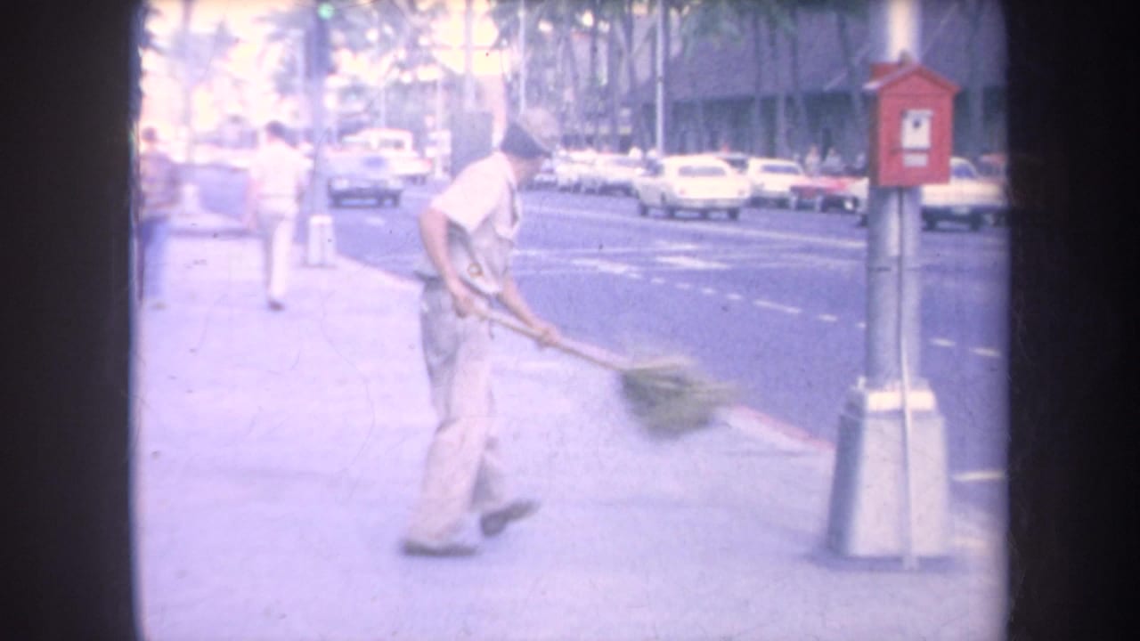 1969 Hawaii home-movie frame showing a man sweeping a sidewalk with a large broom near traffic, palm trees, and a red utility box.