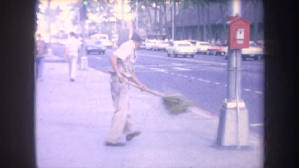 1969 Hawaii home-movie frame showing a man sweeping a sidewalk with a large broom near traffic, palm trees, and a red utility box.