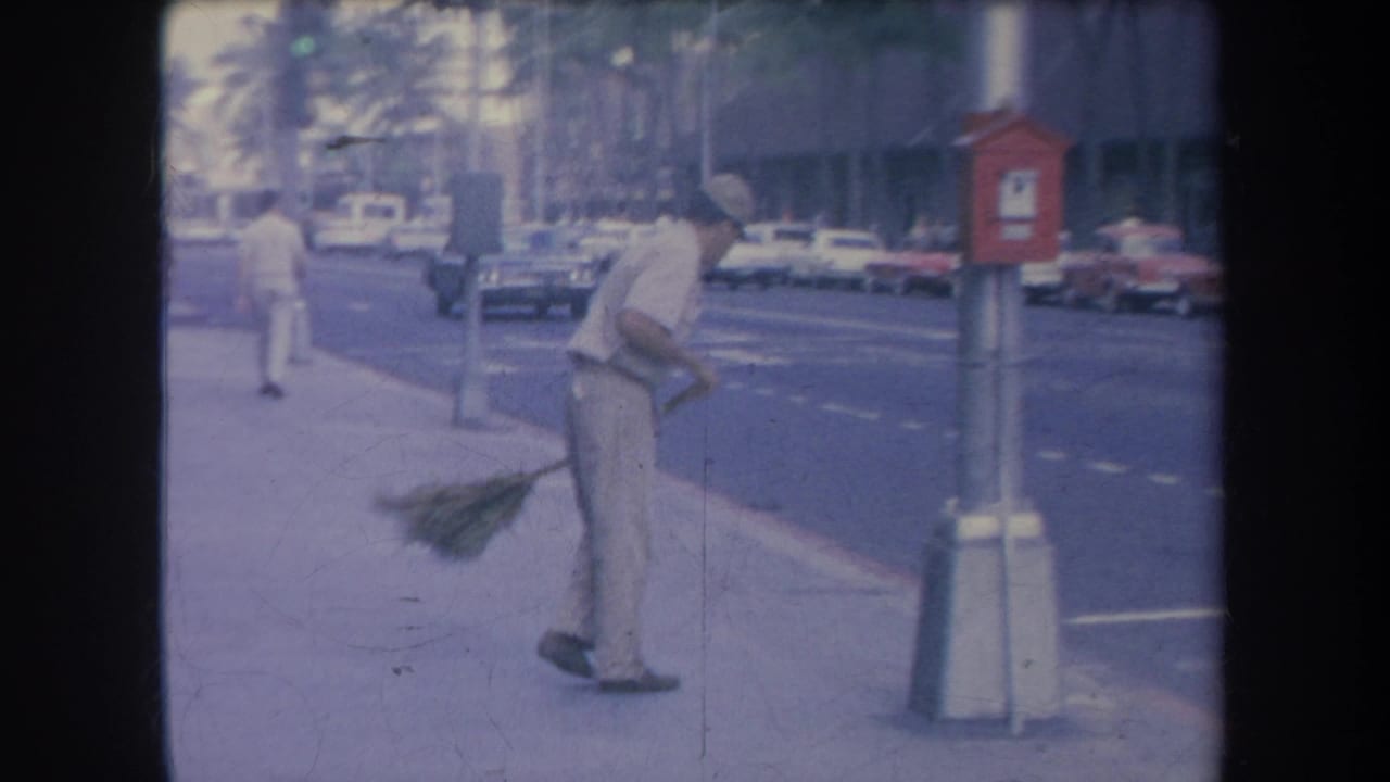 1969 Hawaii street frame with a uniformed man sweeping near the curb while cars and palm trees appear along the road.