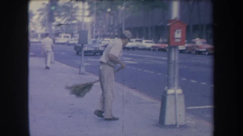 1969 Hawaii street frame with a uniformed man sweeping near the curb while cars and palm trees appear along the road.