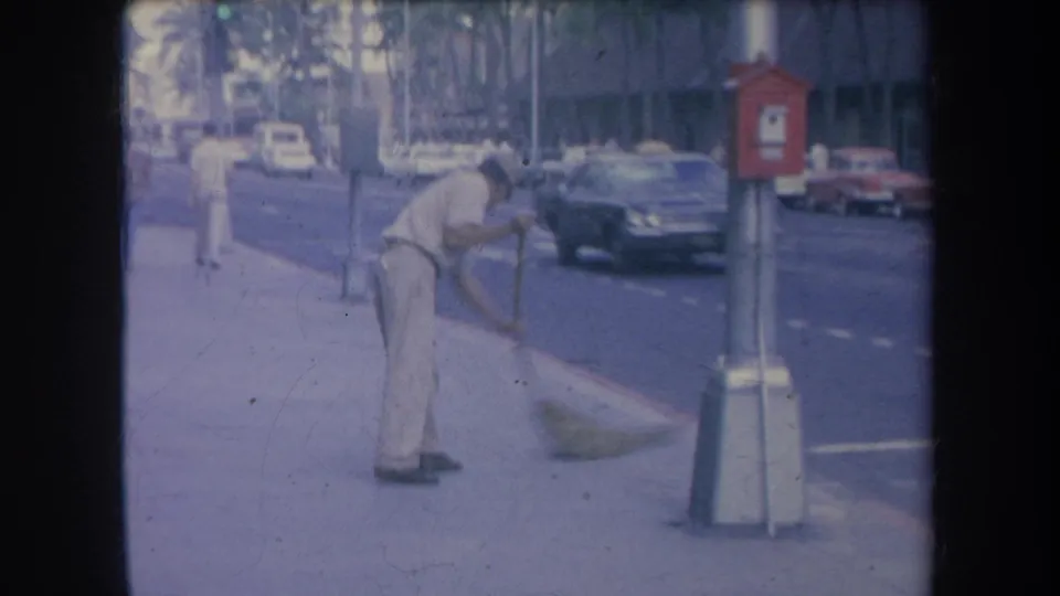 1969 Hawaii home-movie frame showing a man sweeping the sidewalk beside passing traffic and a red sidewalk box.
