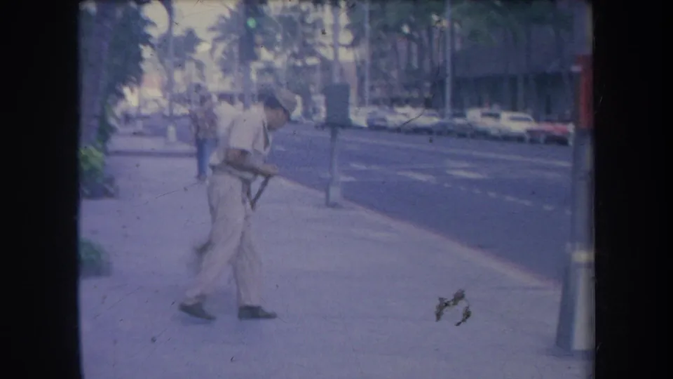 1969 Hawaii frame showing the sidewalk worker carrying or sweeping with a large broom as pedestrians and cars remain in the background.