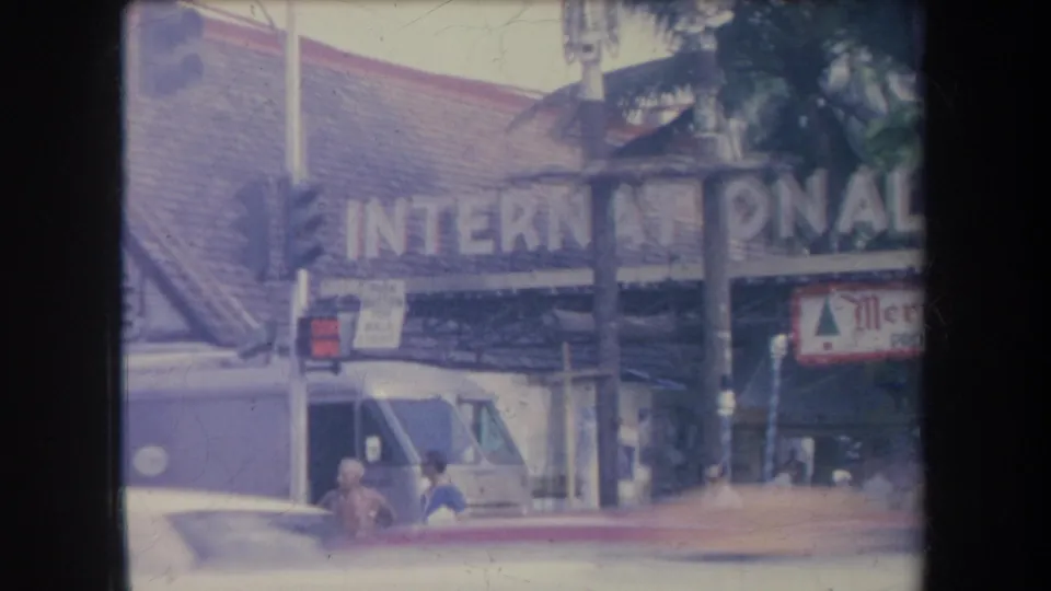 1969 Hawaii home-movie frame showing the International Market sign, a white van, pedestrians, and a Merry Christmas banner.