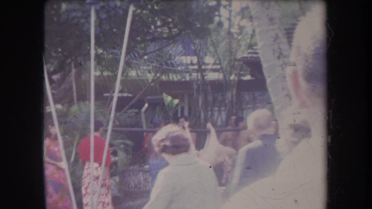 1969 Hawaii 8mm frame showing onlookers near palm trees and thatched roofs during an outdoor gathering.