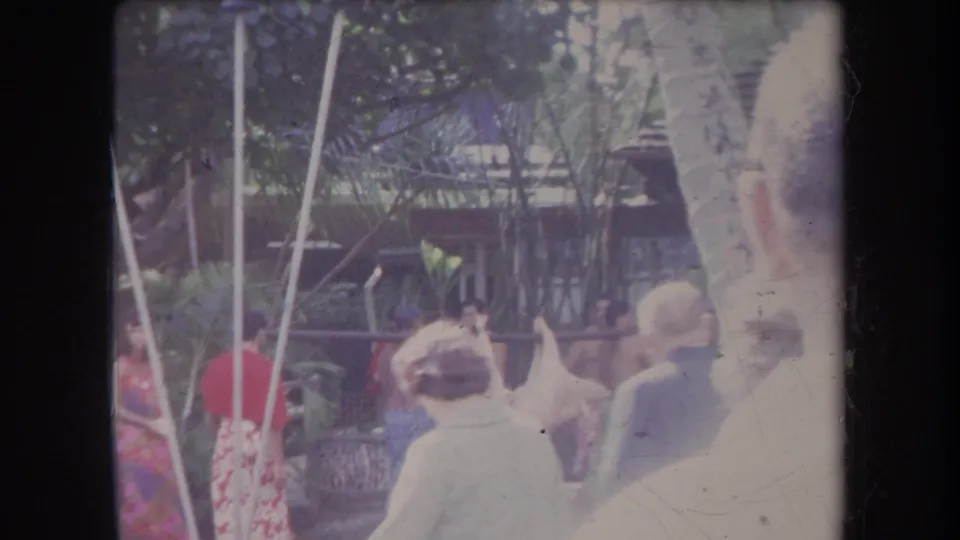1969 Hawaii 8mm frame showing onlookers near palm trees and thatched roofs during an outdoor gathering.