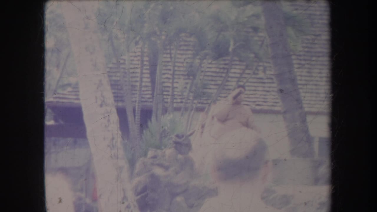 A shirtless man in a light wrap stands near tropical plants as people watch from nearby.