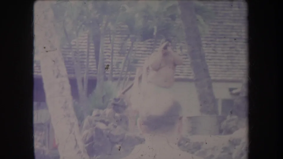 Faded 1969 Hawaii 8mm frame showing a man in a light wrap near onlookers, palms, and thatched-roof structures.