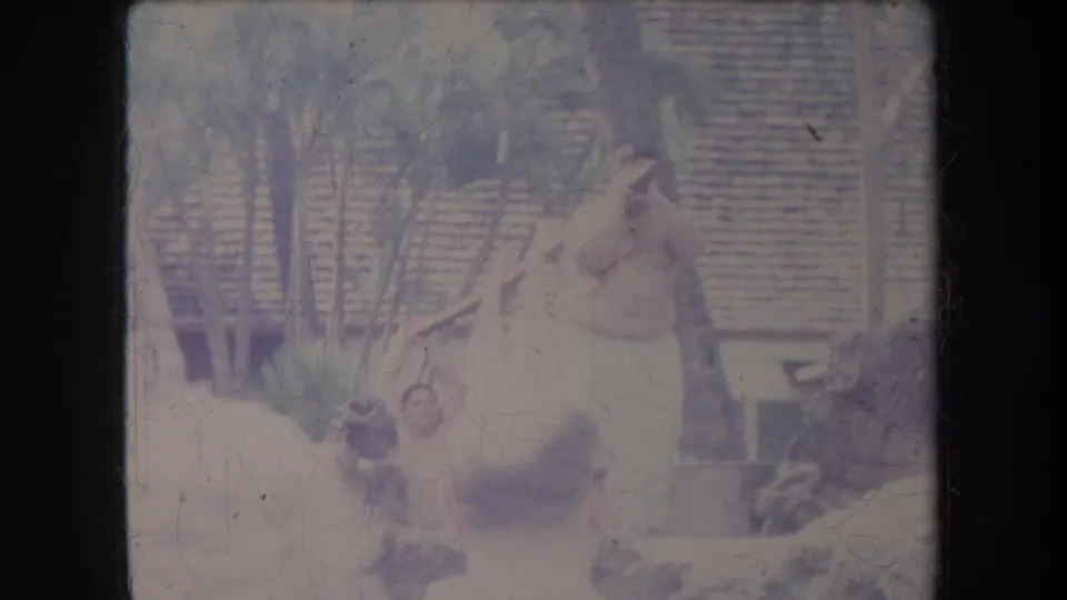 1969 Hawaii 8mm frame of a shirtless man in a light wrap near palms and thatched roofing, seen past spectators.
