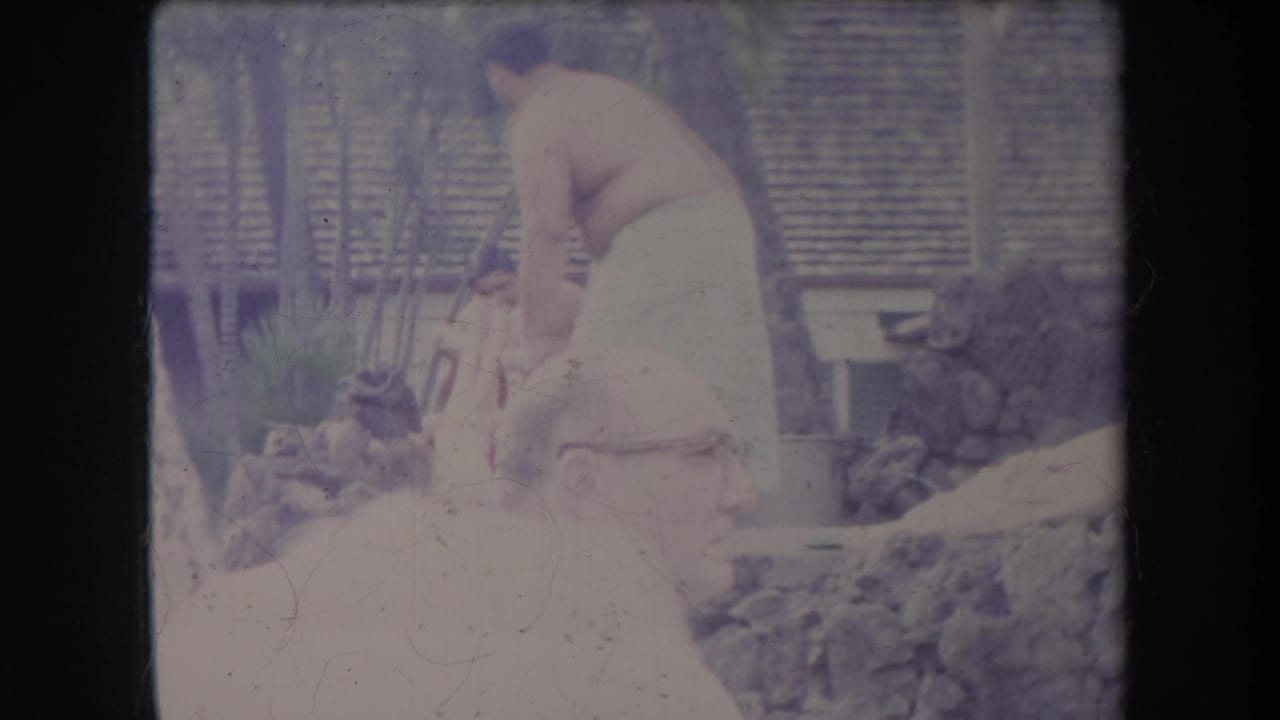 1969 Hawaii home-movie frame showing a man bending over a low fire or cooking pit as another man stands nearby and onlookers sit in the foreground.