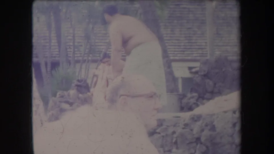 1969 Hawaii home-movie frame showing a man bending over a low fire or cooking pit as another man stands nearby and onlookers sit in the foreground.