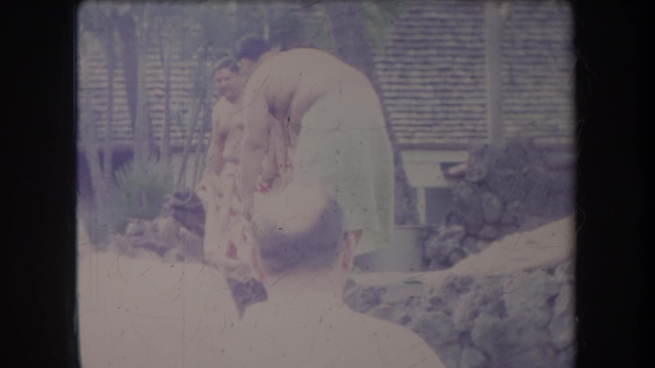 Standing man watches another man bend over a low outdoor cooking area.
