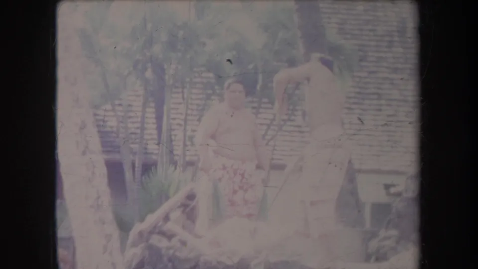 Faded 1969 Hawaii home-movie frame showing a man in a patterned wrap near a tree, with another man working by the luau cooking area.