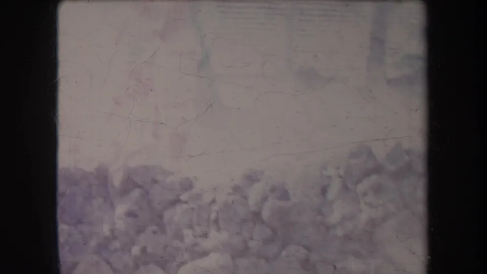 Very faded 1969 Hawaii frame with a barely visible person near the luau area, rocks in the foreground, and tropical foliage or thatch behind.
