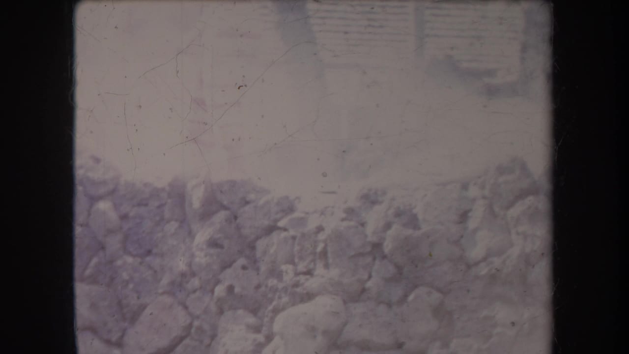 Faded 1969 Hawaii home-movie frame with rocks or a stone wall and a hazy outdoor background, with no clear faces visible.